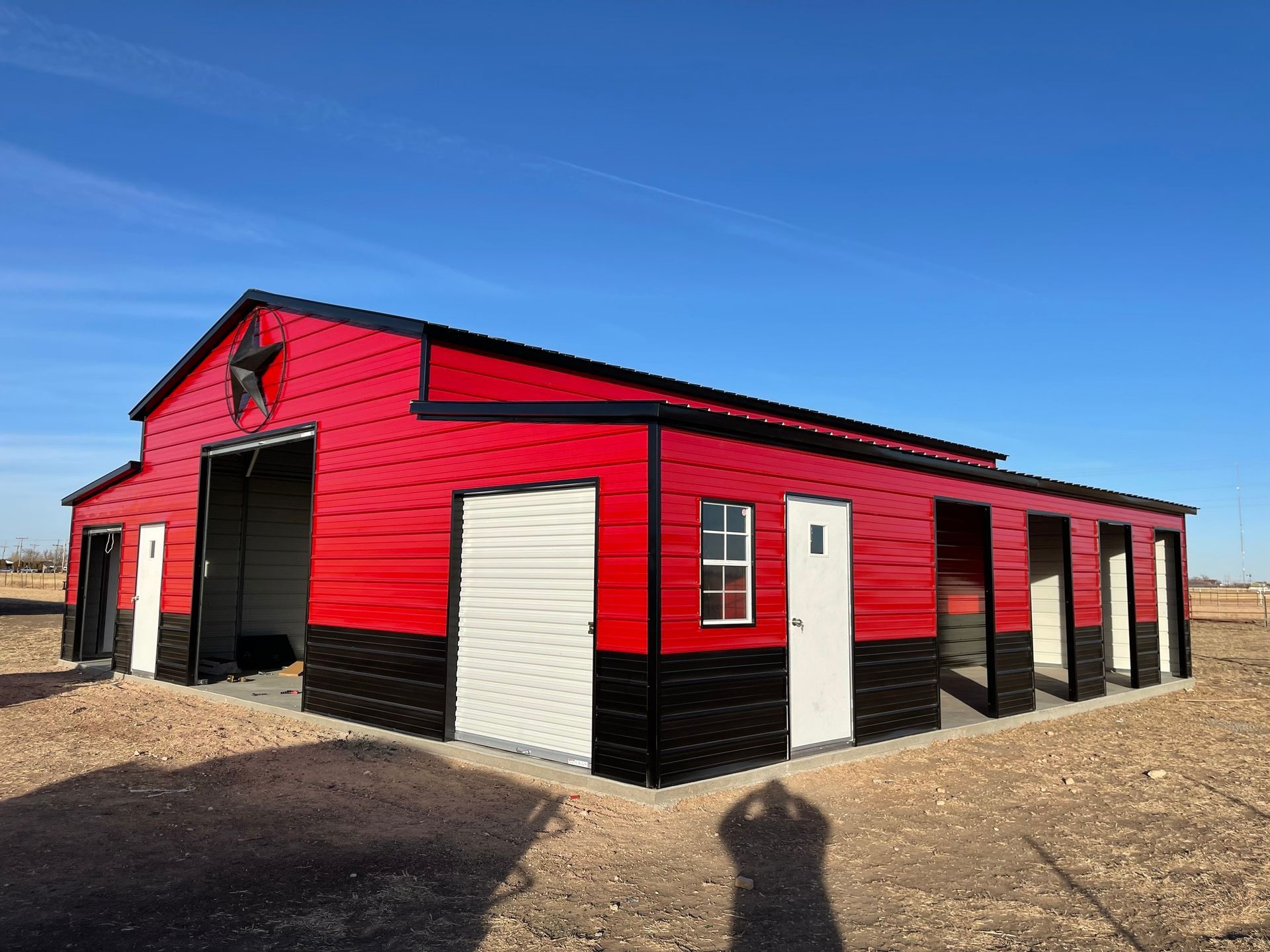 Red and black barn-style metal building with open garage bay, doors, and a star on the gable against a blue sky.