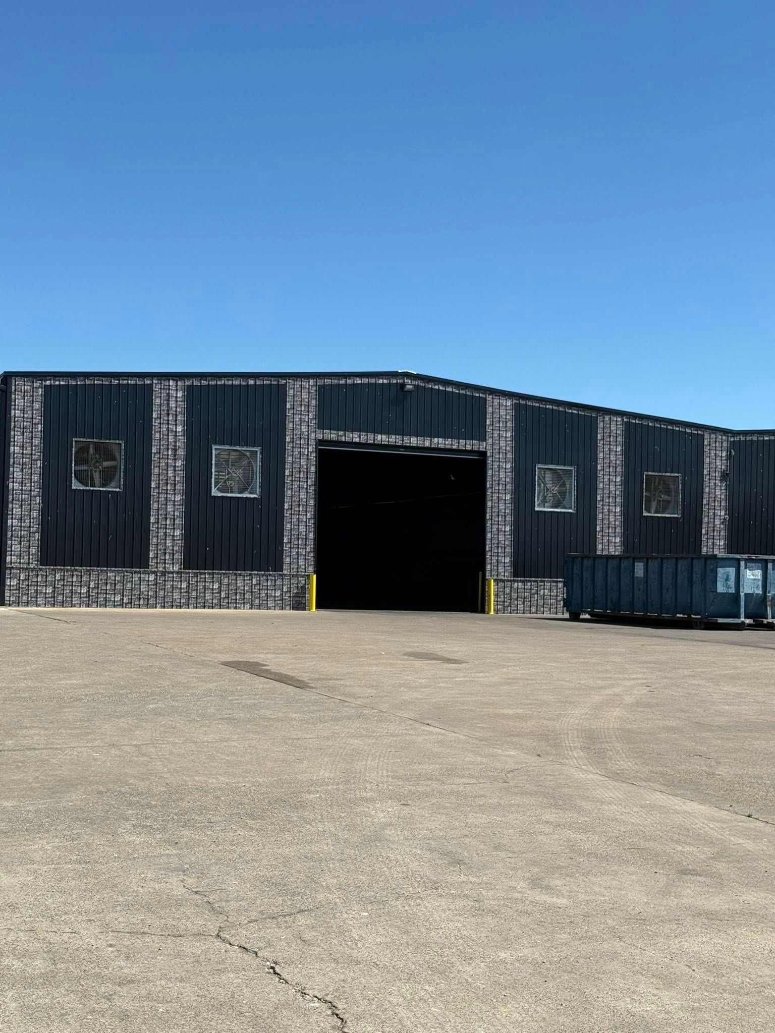 Dark industrial building with large open doorway, surrounded by asphalt and a blue sky.