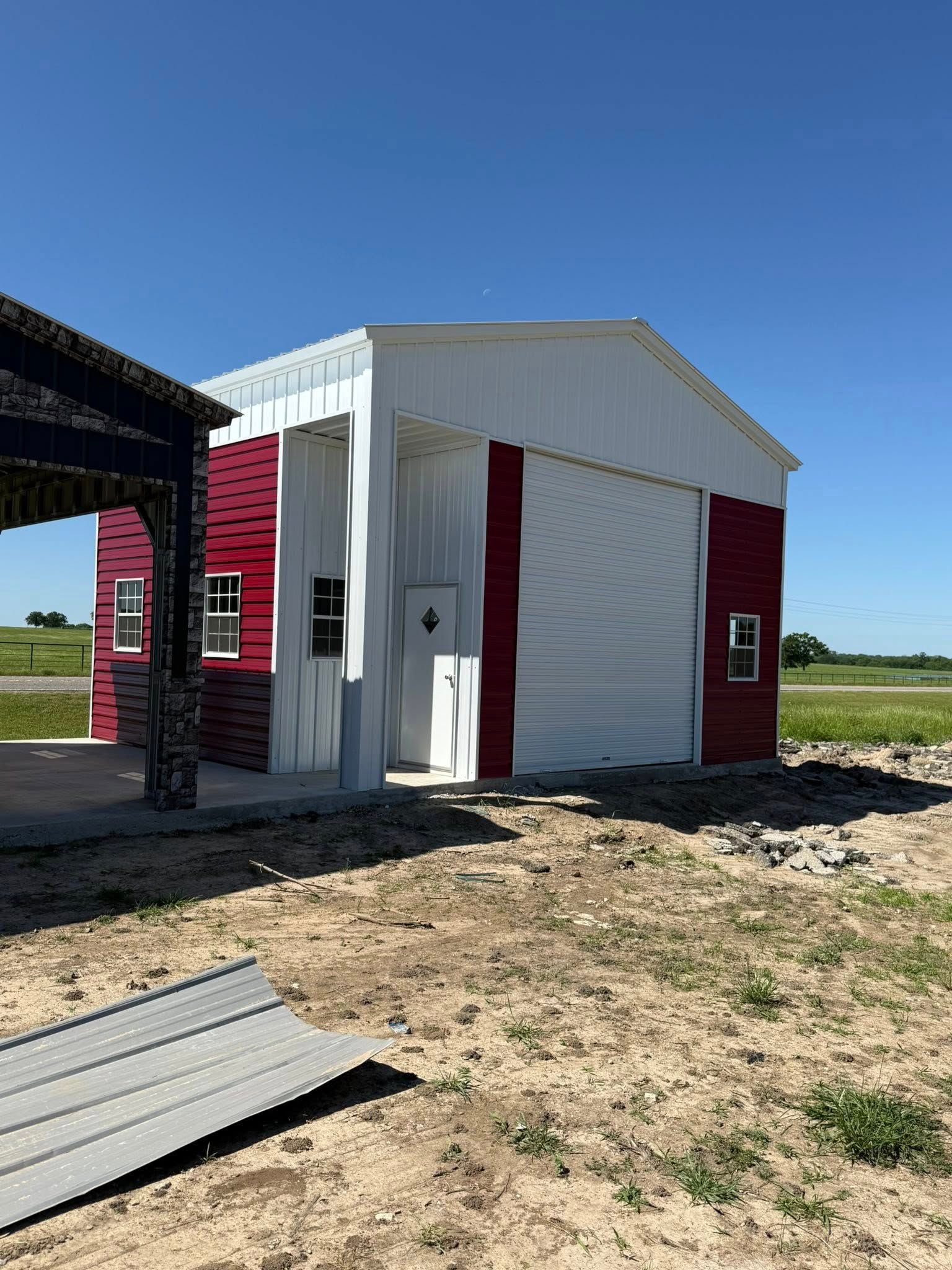 Red and white metal building with a roll-up door, next to a shaded structure, set in a grassy field.