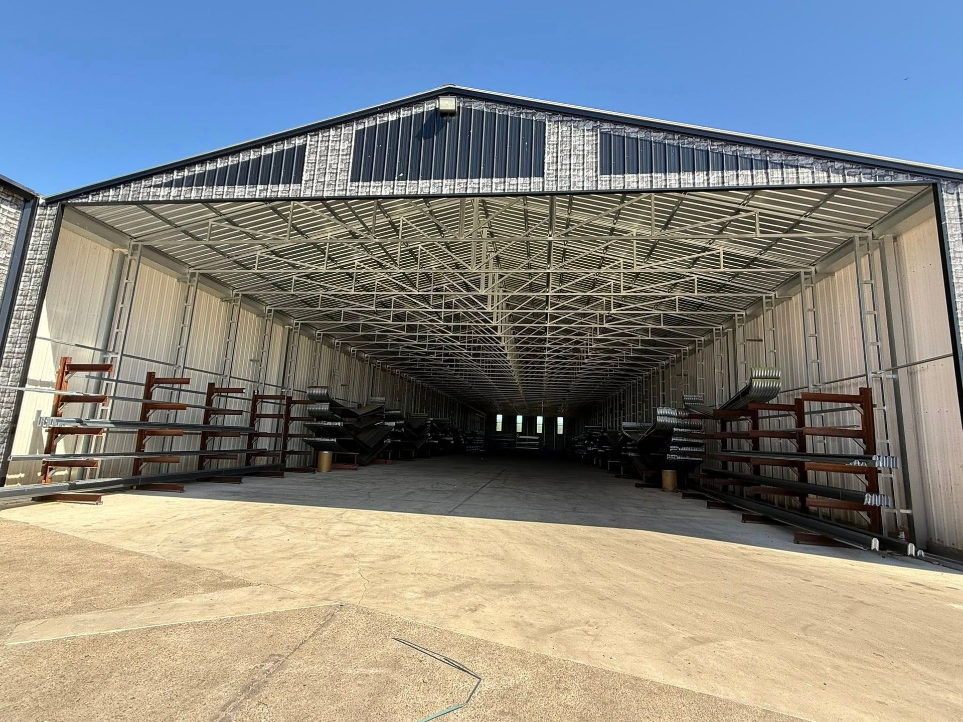 Open-ended metal shed with shelving on sides, a long interior, and a gravel floor under a blue sky.