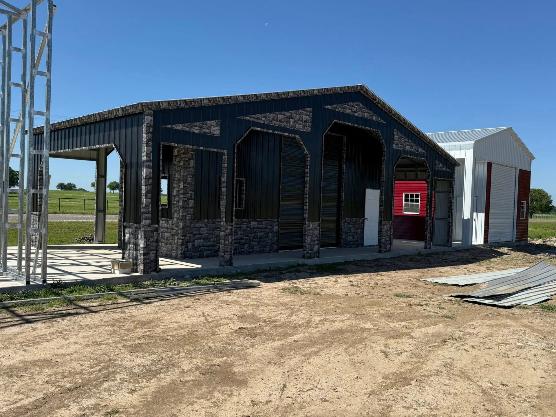 A partially-built black metal barn with stone accents on a sunny day, with a red building and other structures in the background.