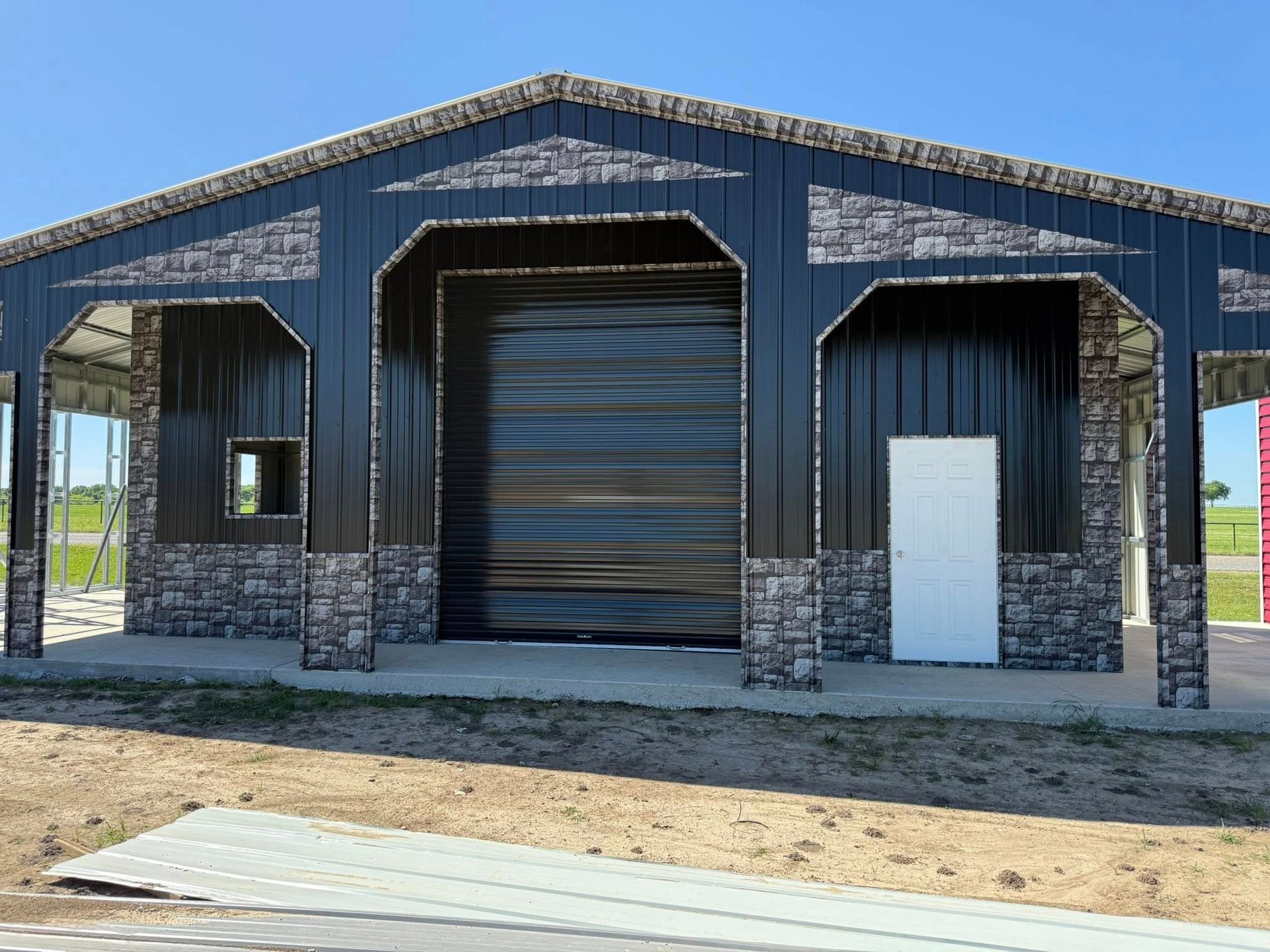 Black metal building with stone accents, large garage door, and white door. Blue sky background.