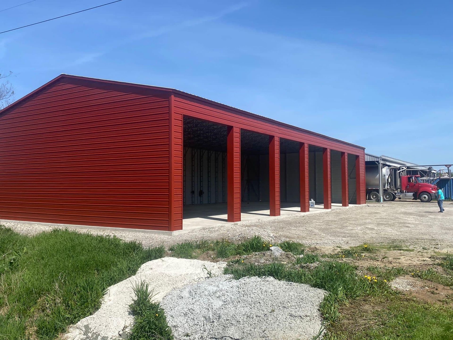 Red metal building with open bays, gravel ground, blue sky.