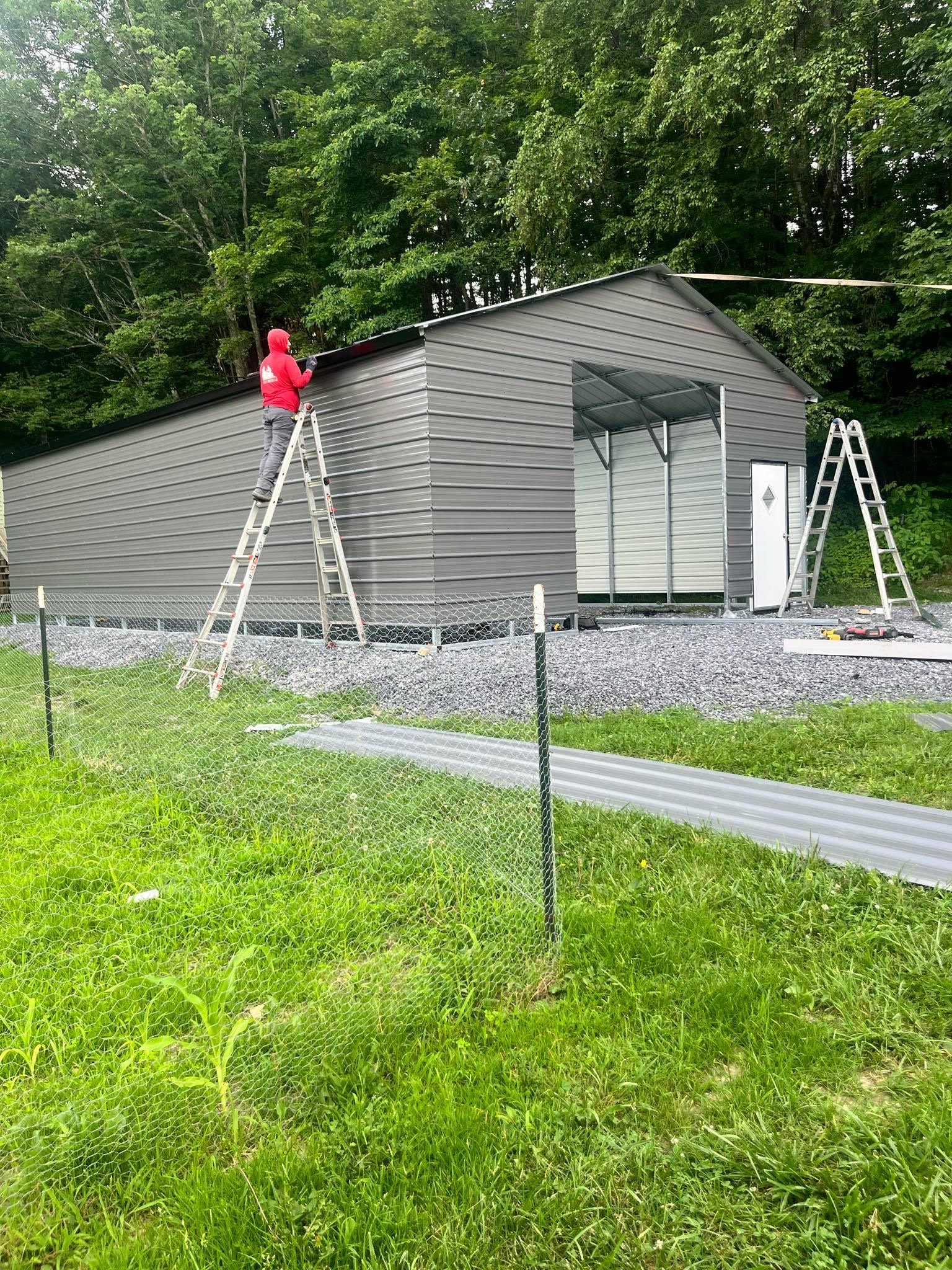 A person on a ladder installing roofing on a grey shed in a grassy field.