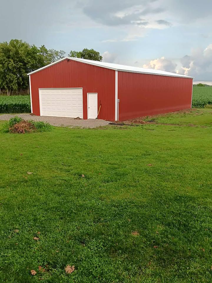 Red metal barn with white garage door and small door, in a grassy field under a cloudy sky.
