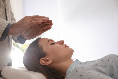 Person receiving Reiki, hands hovering above head, eyes closed. Light-filled room.