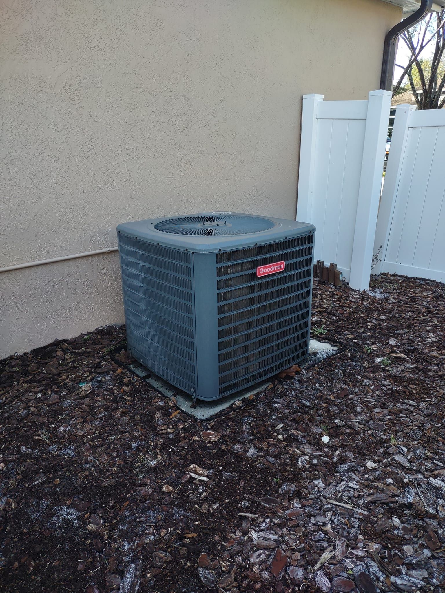 a gray air conditioner is sitting on top of a pile of mulch next to a white fence .