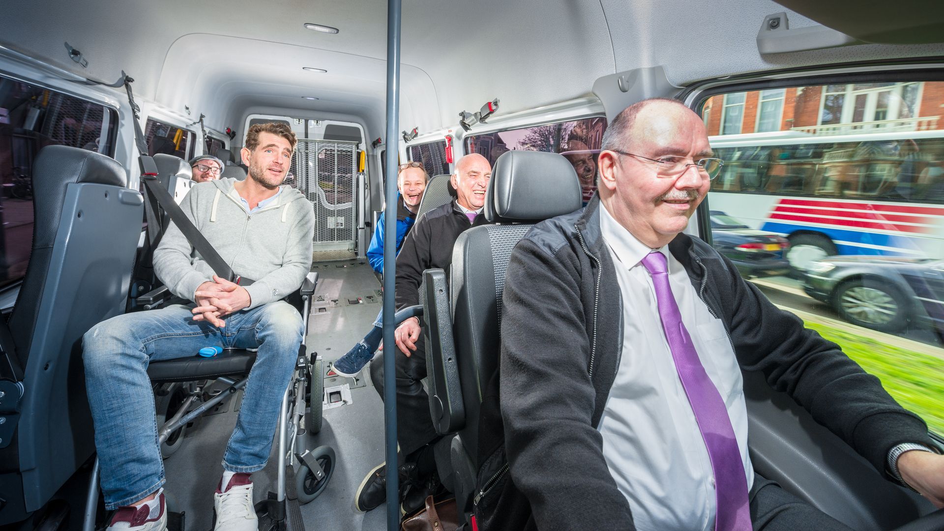 A group of people travelling in a taxi with the driver and a person in a wheelchair upfront.