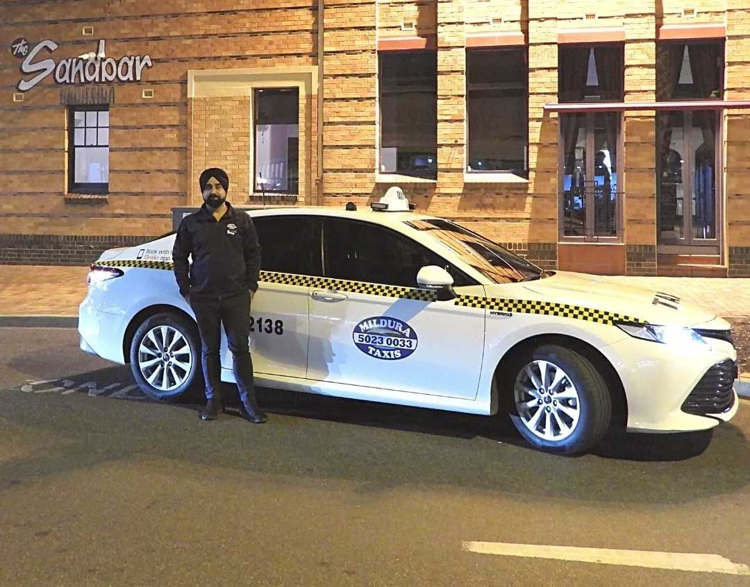 Yellow taxi sign atop a white car, near a building and outdoor seating area.