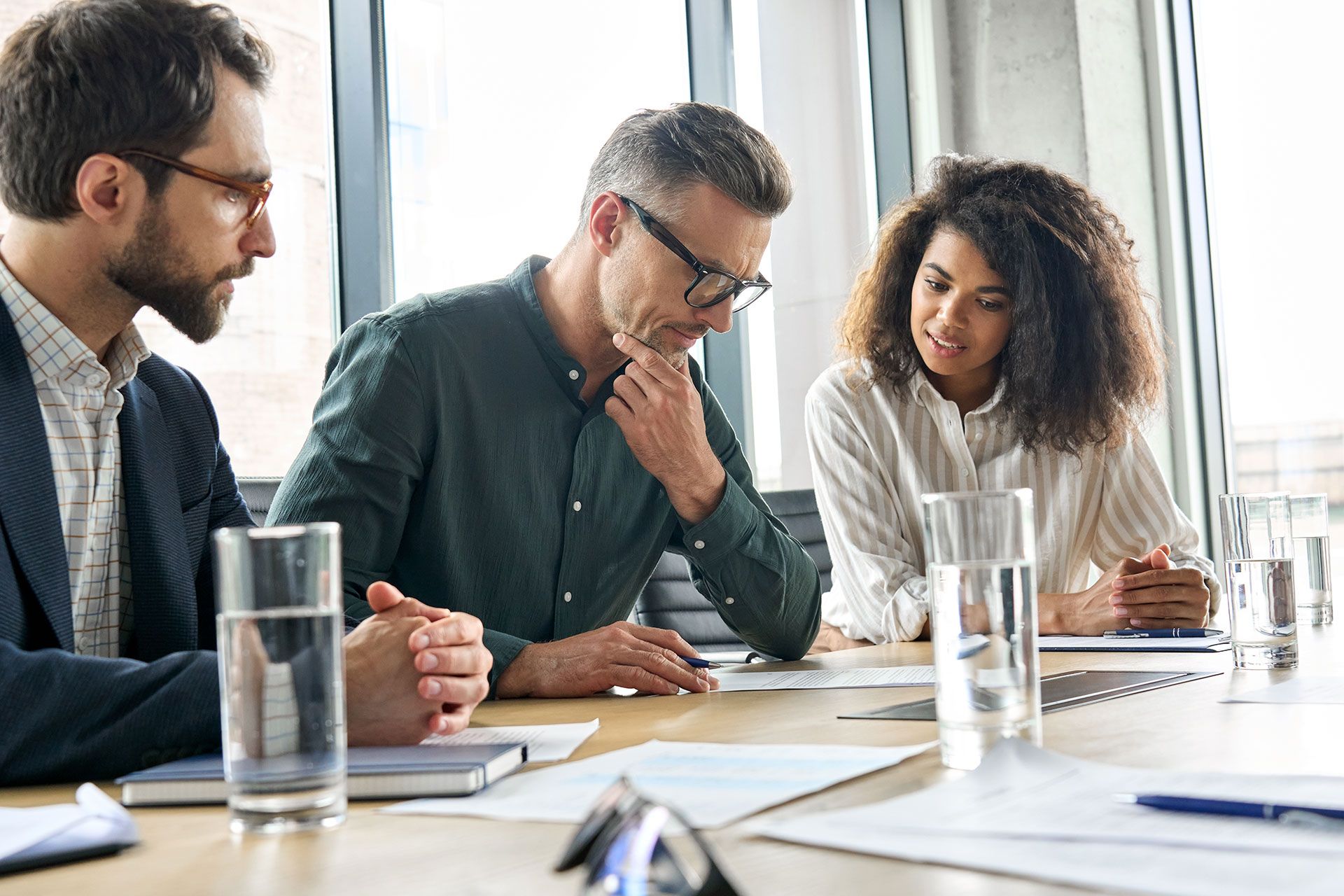 Three people in a meeting, looking at papers on a table. The man in the middle is deep in thought.