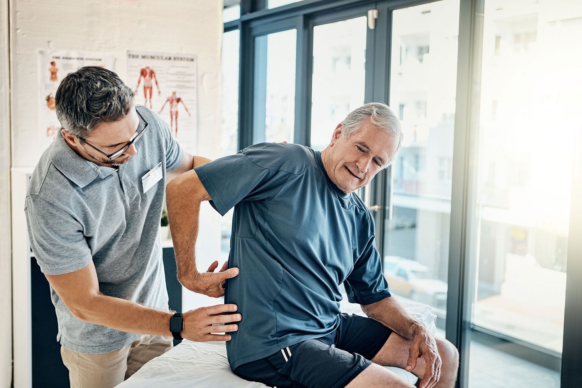 Physical therapist examining a man's lower back; man smiles, leaning to the side. Clinic setting.