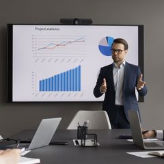 Man presenting charts on screen in a meeting, gesturing with hands, wearing a suit.