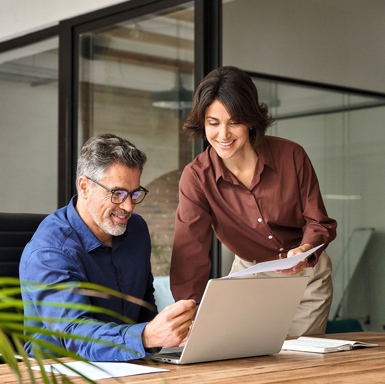 Man and woman looking at laptop in office, woman holding papers, smiling.