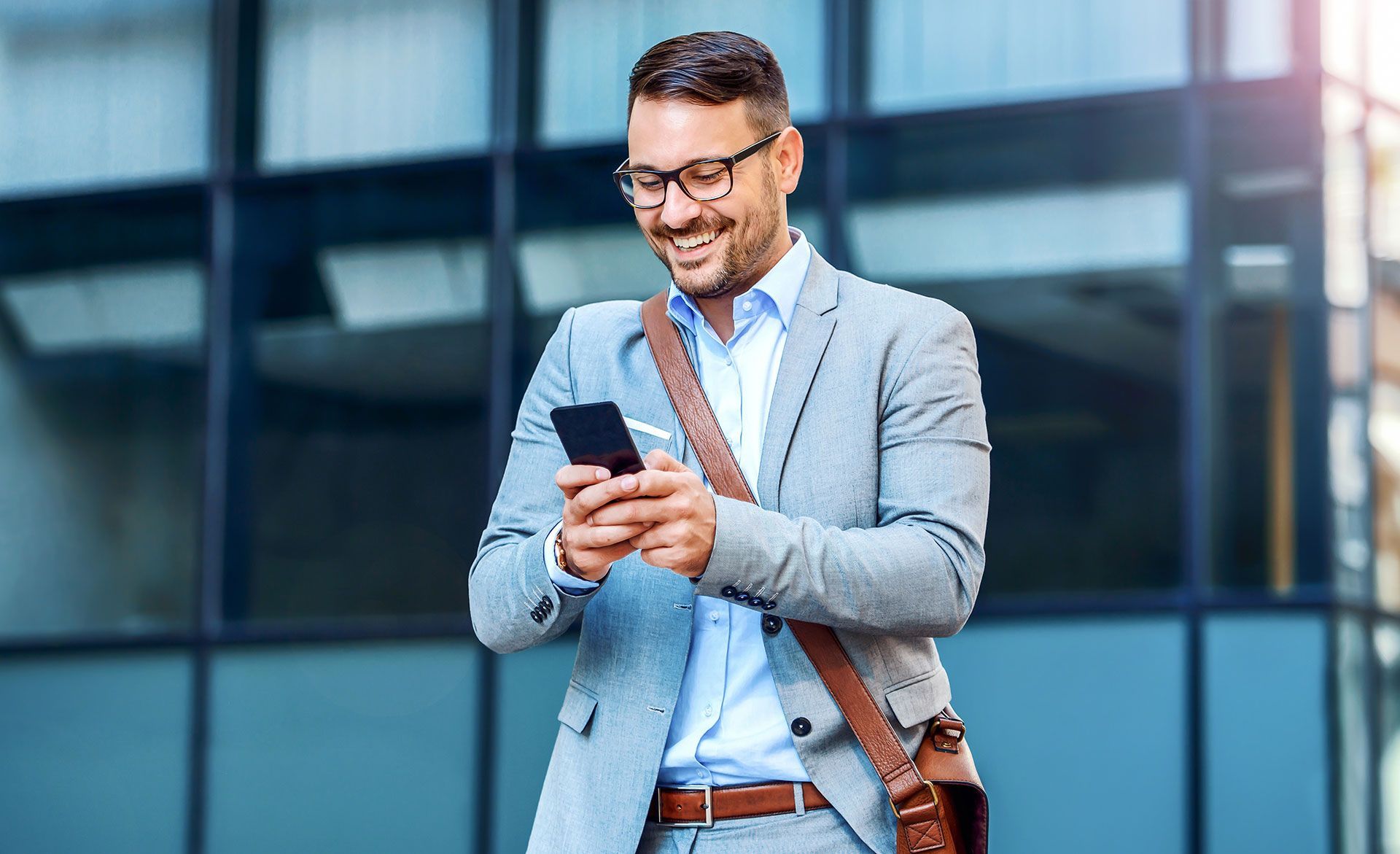 Man in suit smiling while looking at his phone outside a modern building.