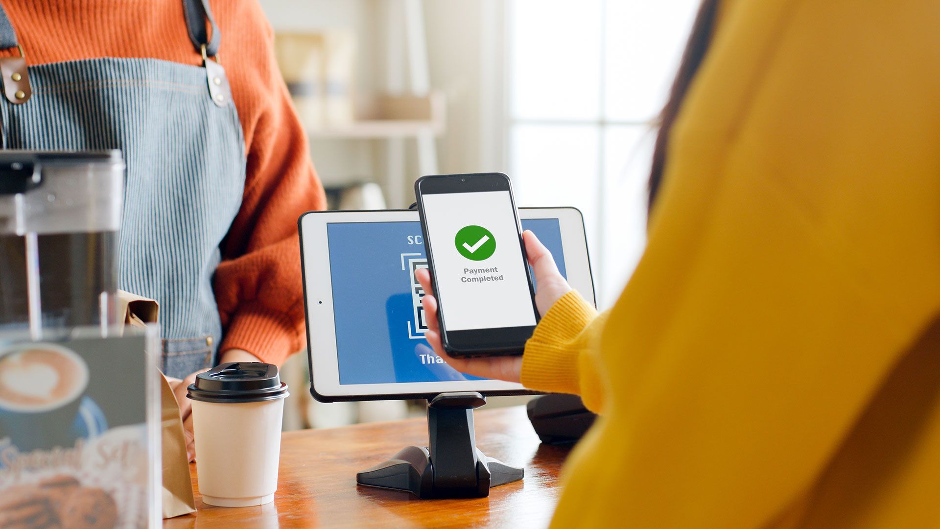 Woman paying with phone at a cafe; phone shows successful payment. Counter, barista, coffee cup in view.