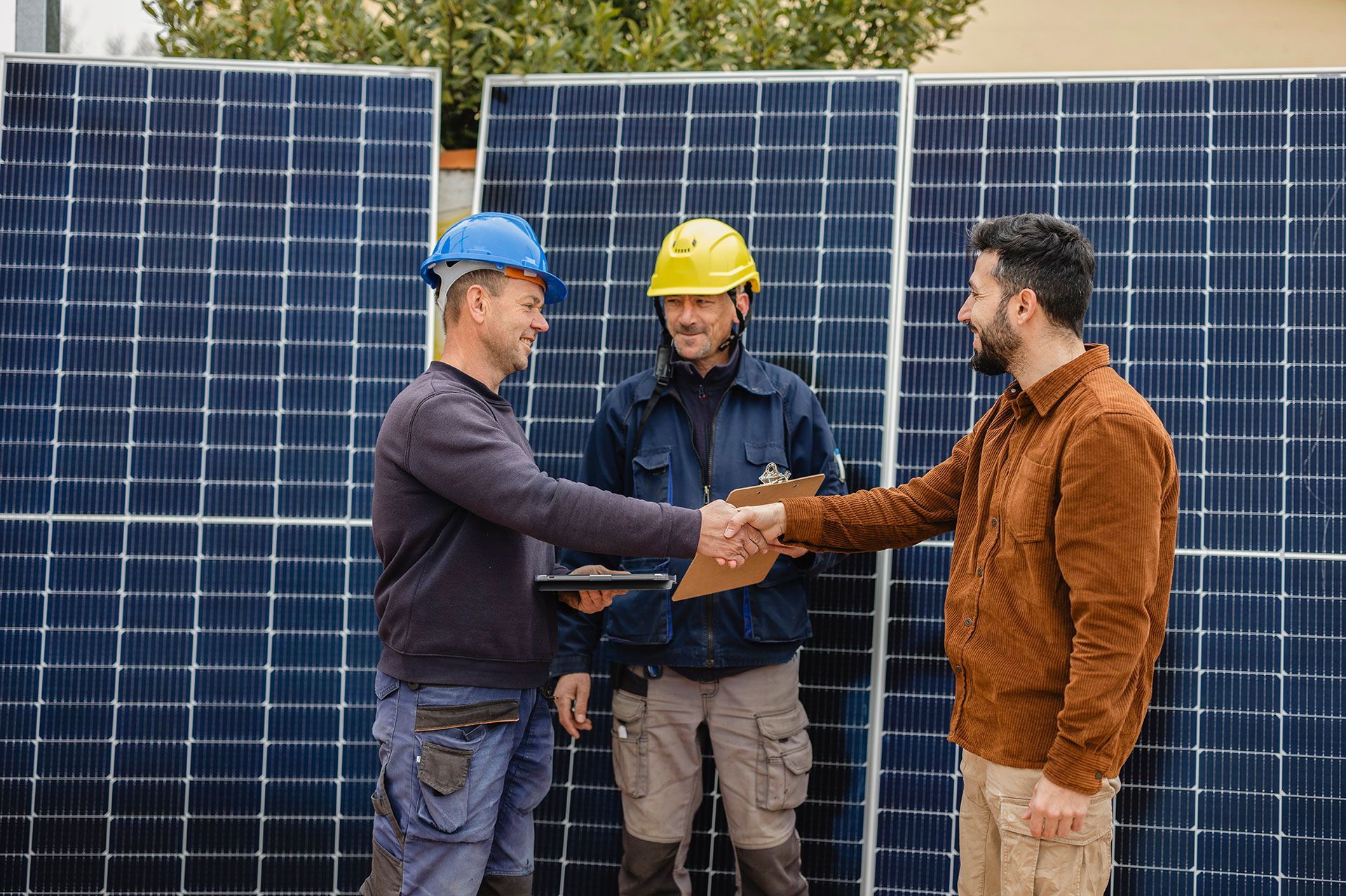 Three men shaking hands in front of solar panels; one wears a hard hat and dark clothes.