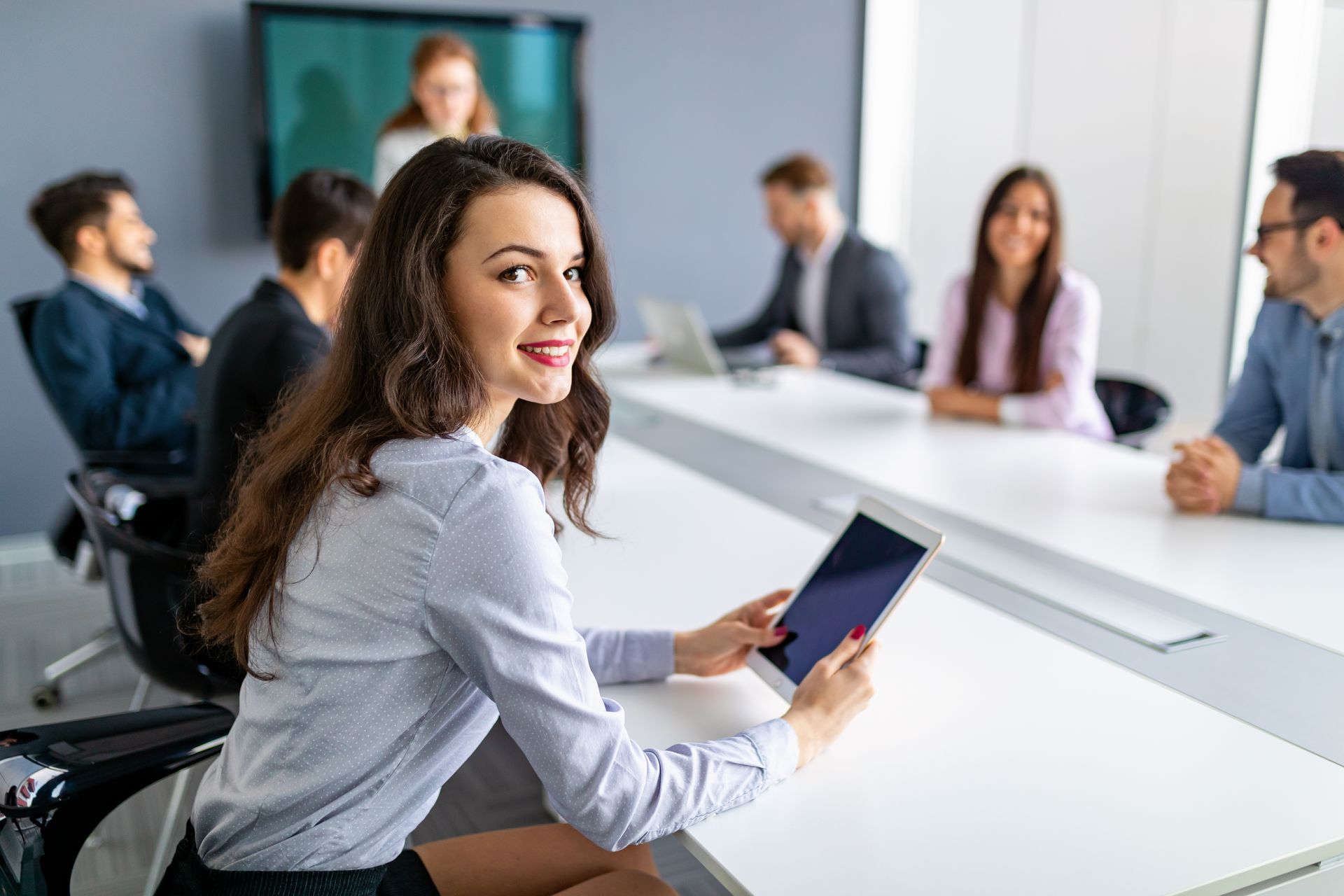 Woman smiles holding tablet in meeting, others around table in modern office.