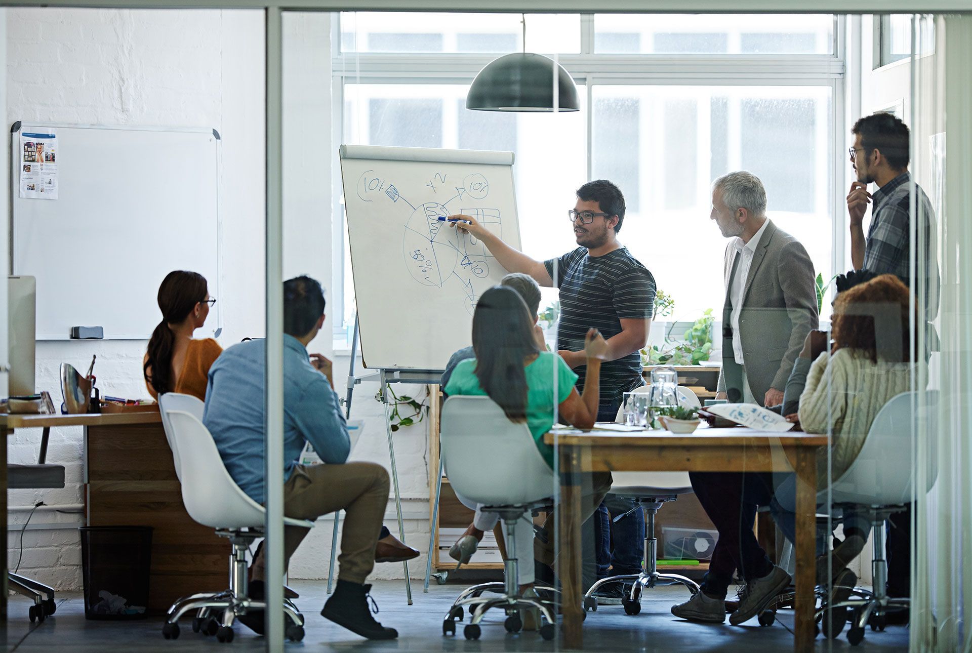 Group of diverse people in a modern office, brainstorming around a whiteboard; one person points.