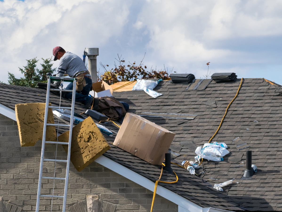 A worker on a ladder performs repairs on a shingled roof with insulation materials and debris scattered nearby.
