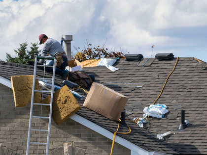 A worker on a ladder performs repairs on a shingled roof with insulation materials and debris scattered nearby.