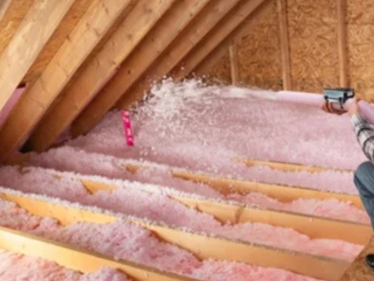 A person uses a hose to blow pink, loose-fill fiberglass insulation into the joist spaces of an unfinished attic.