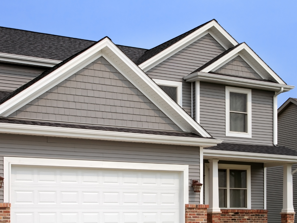 A suburban two-story house exterior featuring gray siding, a white garage door, and a brick base under a clear blue sky.