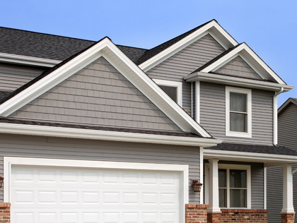 A suburban two-story house exterior featuring gray siding, a white garage door, and a brick base under a clear blue sky.