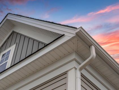 The corner of a white house with gray vertical siding and a gutter under a colorful sunset sky.