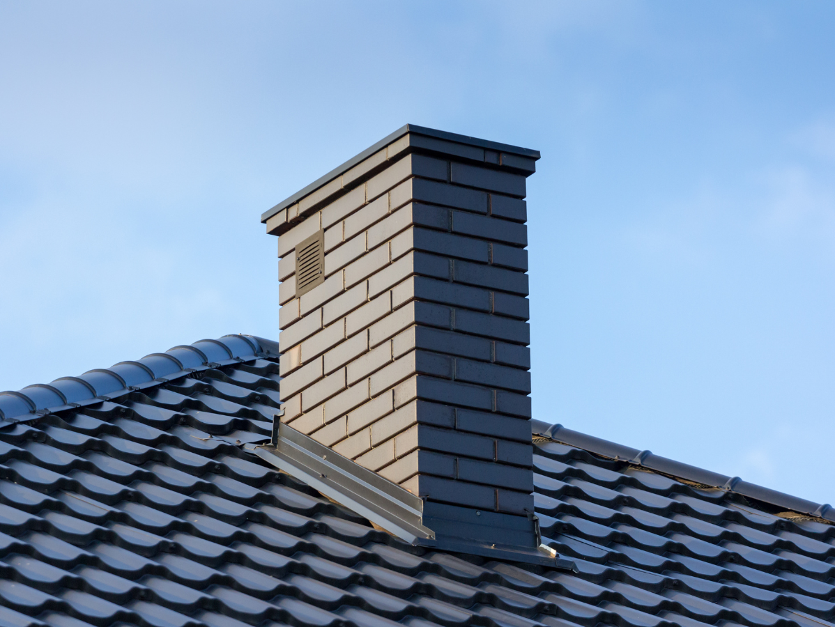 A dark-tiled chimney stands on a pitched roof with matching dark tiles against a clear blue sky.