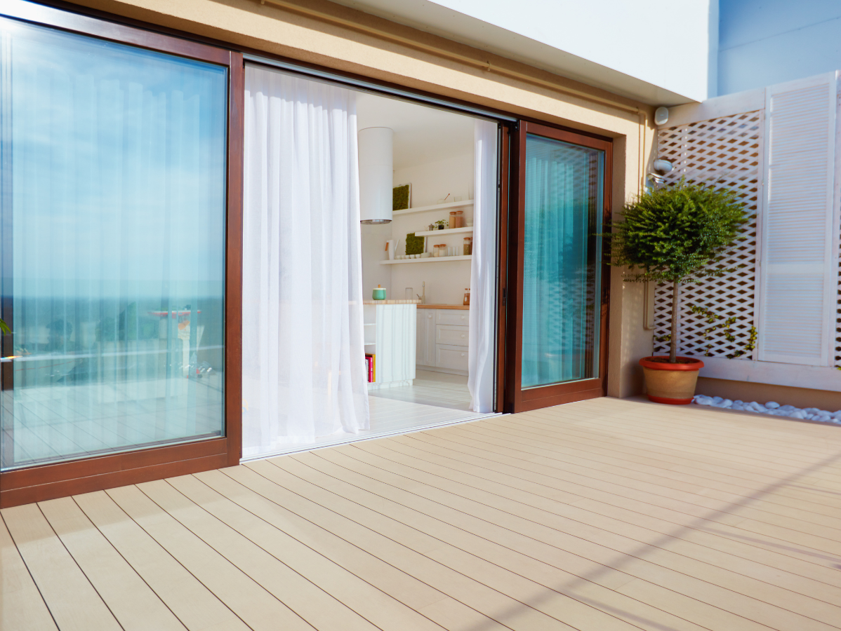 A patio with light-colored decking, large glass sliding doors, white curtains, and a potted tree against a lattice wall.
