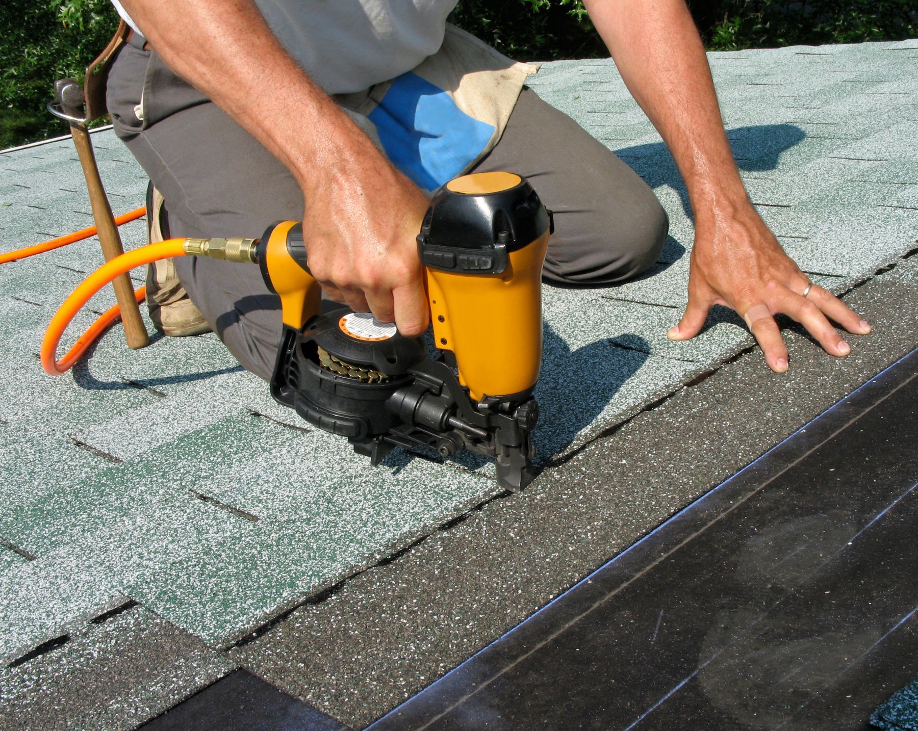 Close-up of a roofing contractor, using a yellow nail gun to apply asphalt shingles to a roof.]