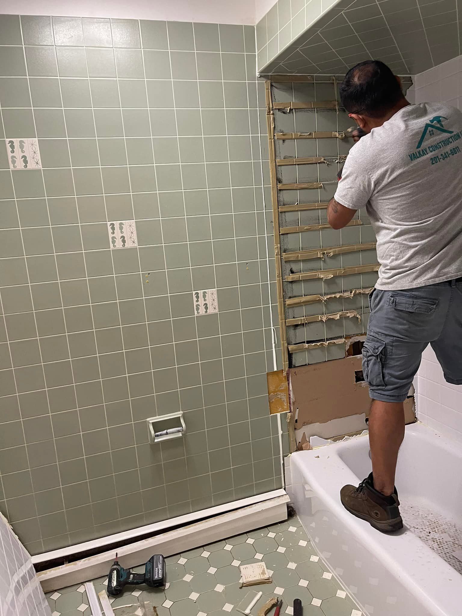 Man removing tiles in bathroom, standing in tub. Green wall, exposed wood, tools, and a white tub are visible.