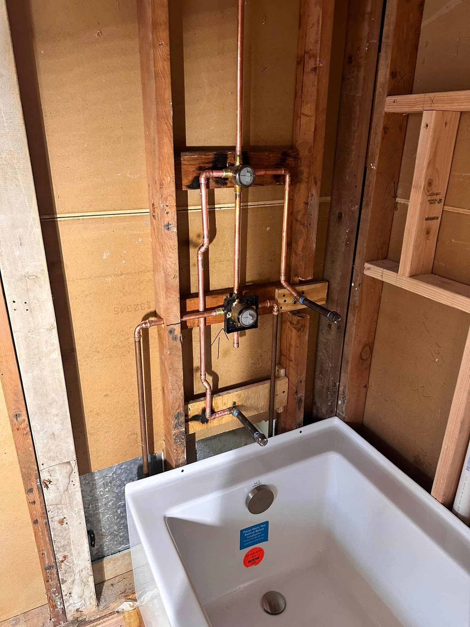 Copper plumbing for a bathtub, installed in a wall with wooden framing, and a white tub below.