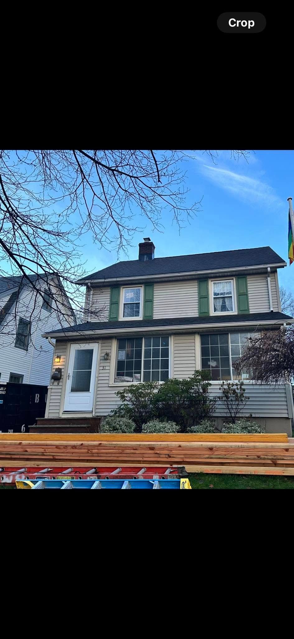 Two-story house with green shutters, white siding, and brown roof. Construction materials stacked in the foreground.