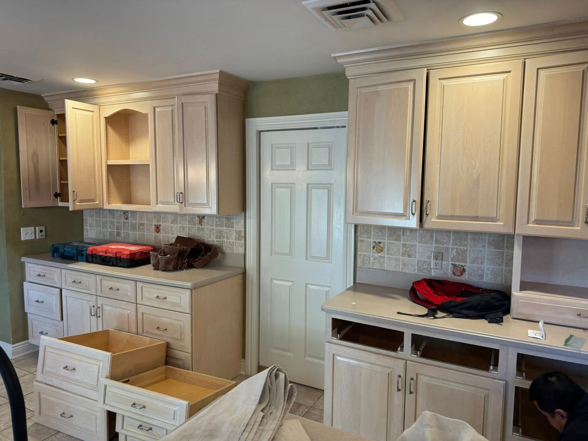 Kitchen cabinets in light color, some open with tools visible. Door in center.