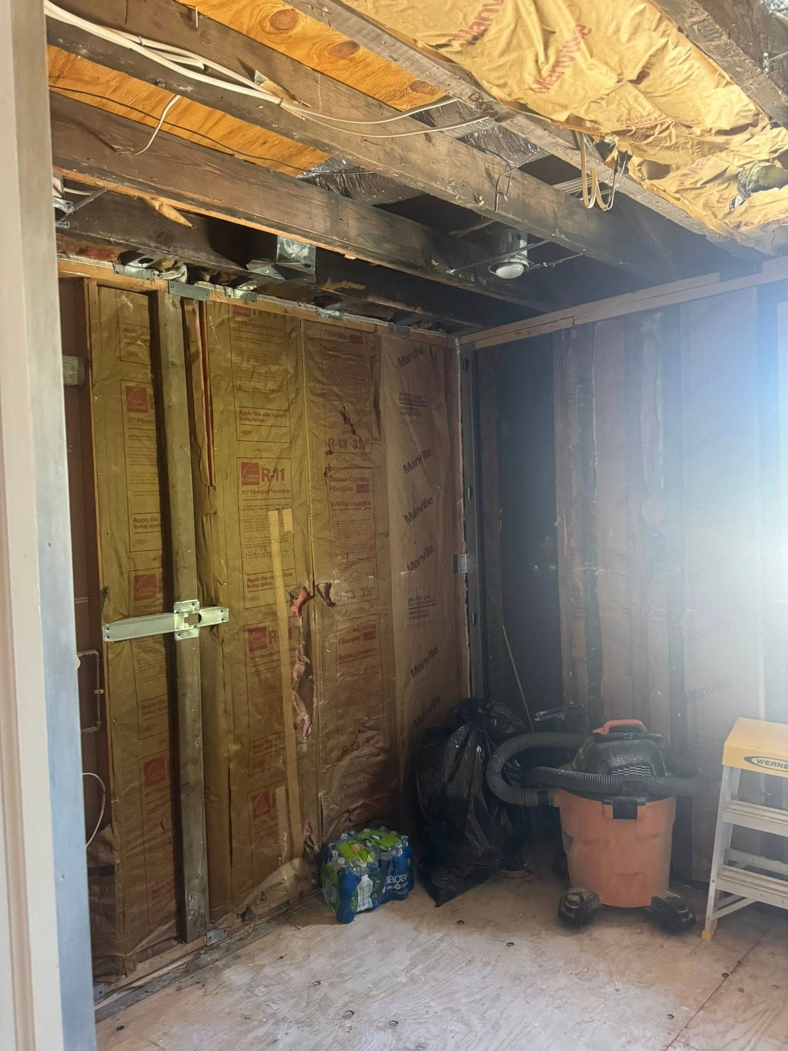 Interior room with damaged walls and ceiling, insulation visible. Debris and tools are present.