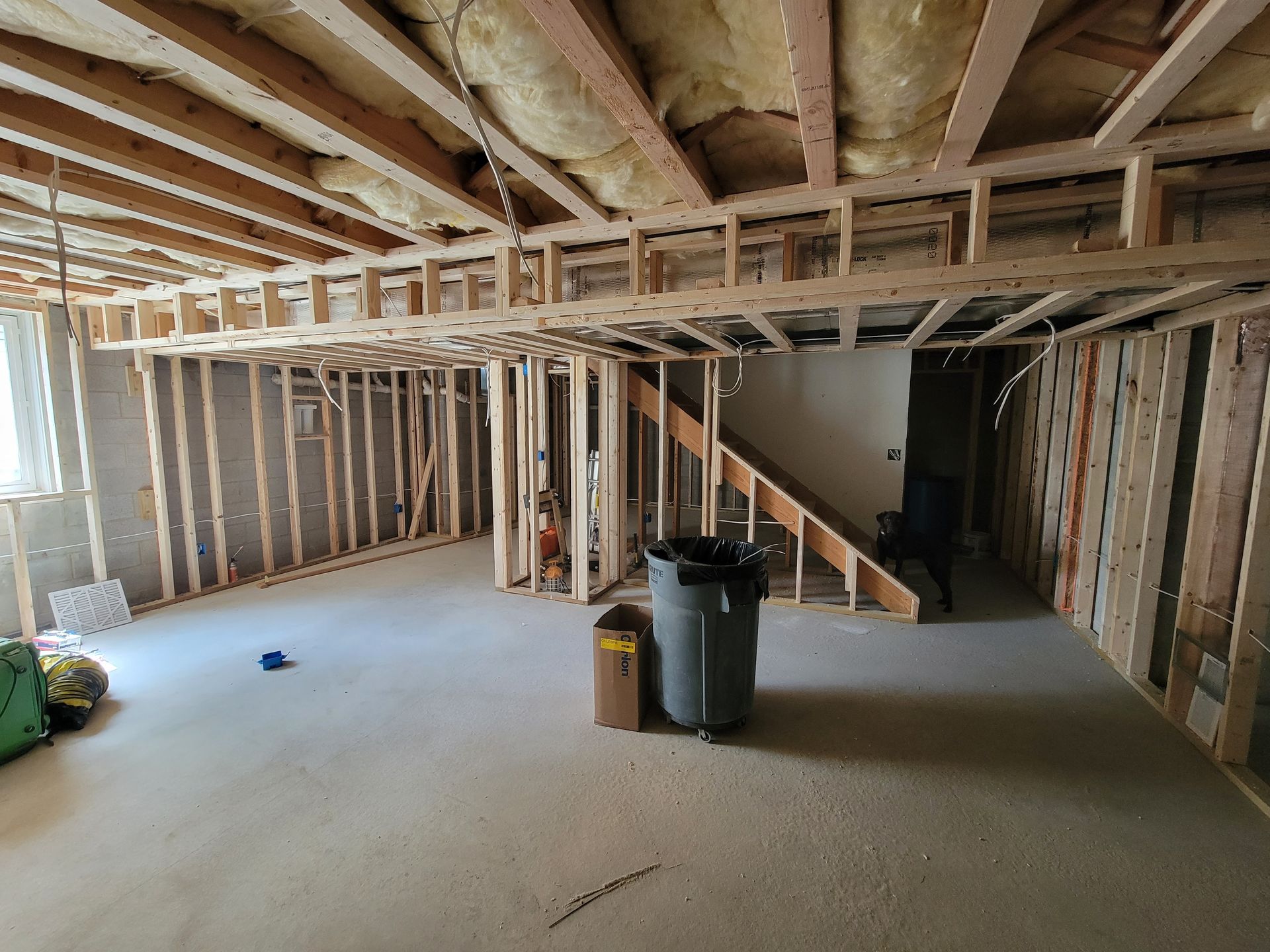 Basement under renovation with exposed wood framing, concrete floor, stairs, and insulation overhead.