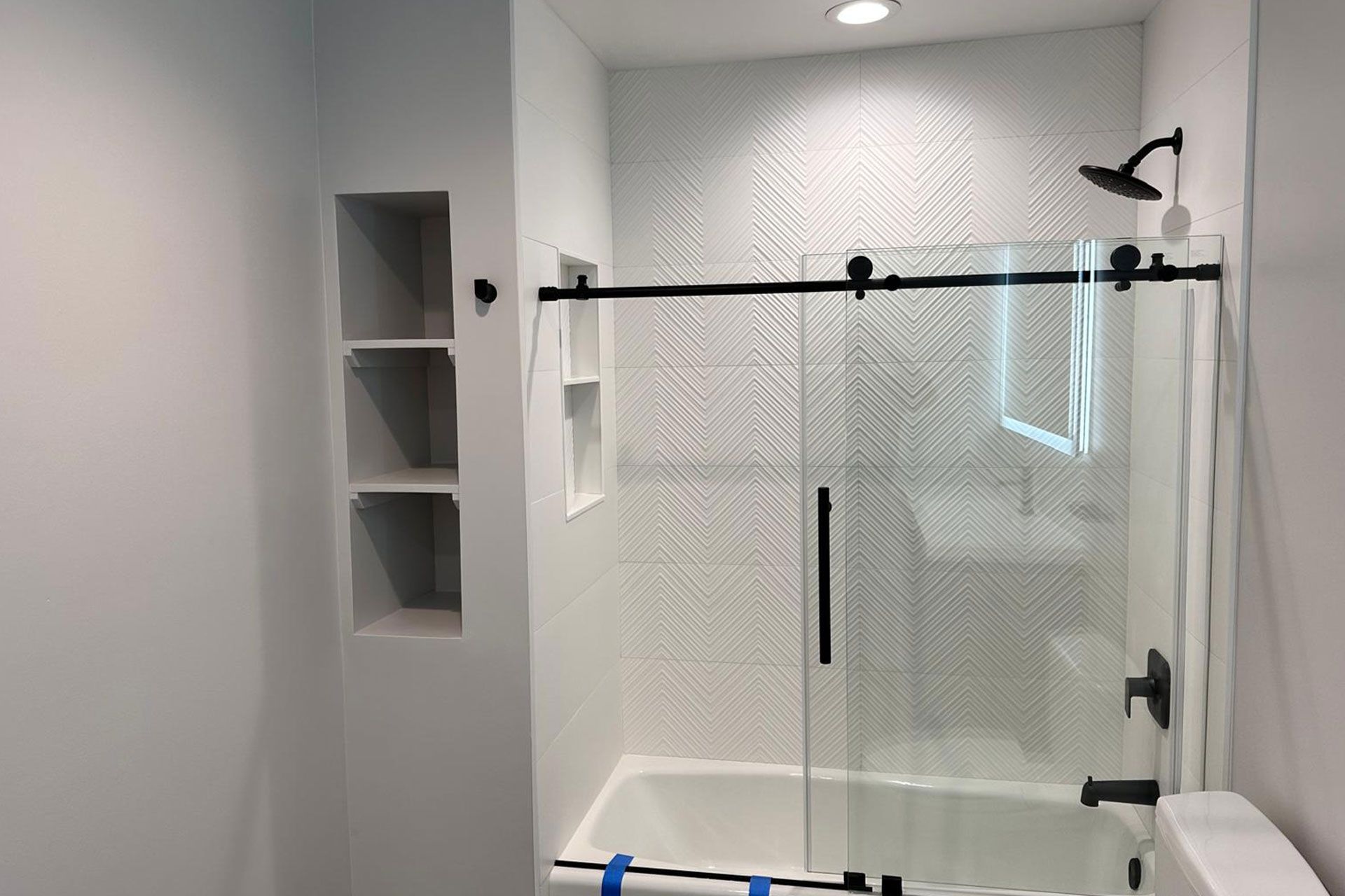 Bathroom with white herringbone tile, glass shower door, black hardware, and recessed shelves.