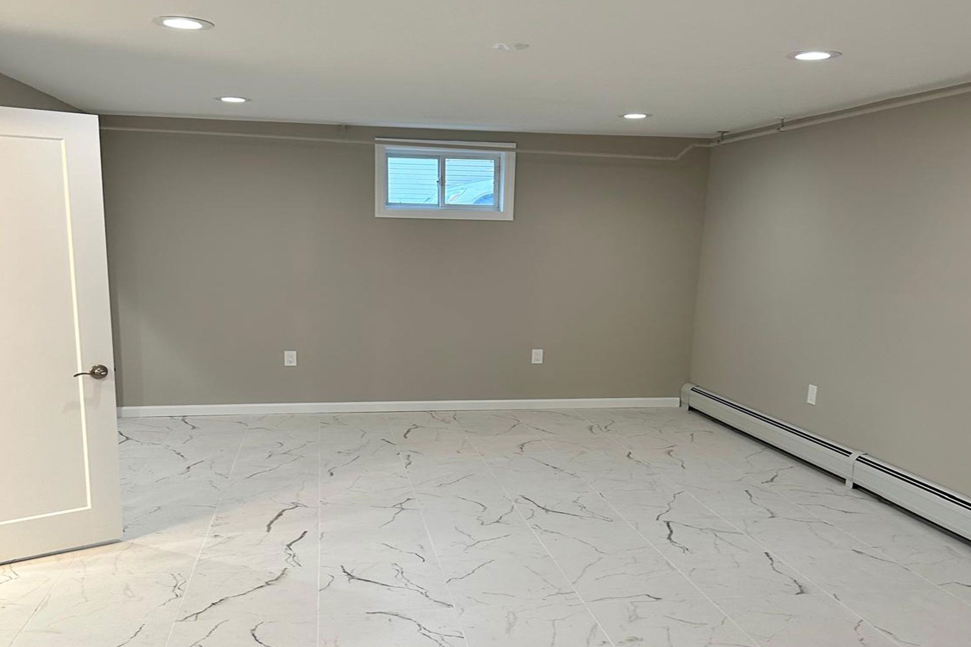 Empty room with light-colored walls and floor, small window, and a radiator.