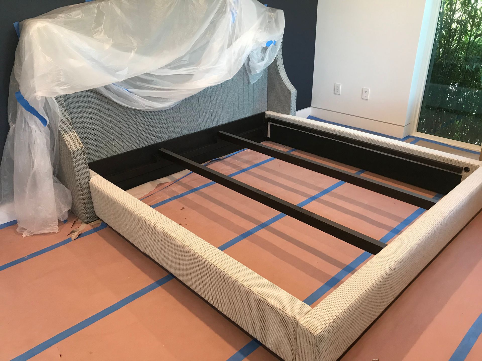A bed frame is sitting on the floor in a room covered in plastic.