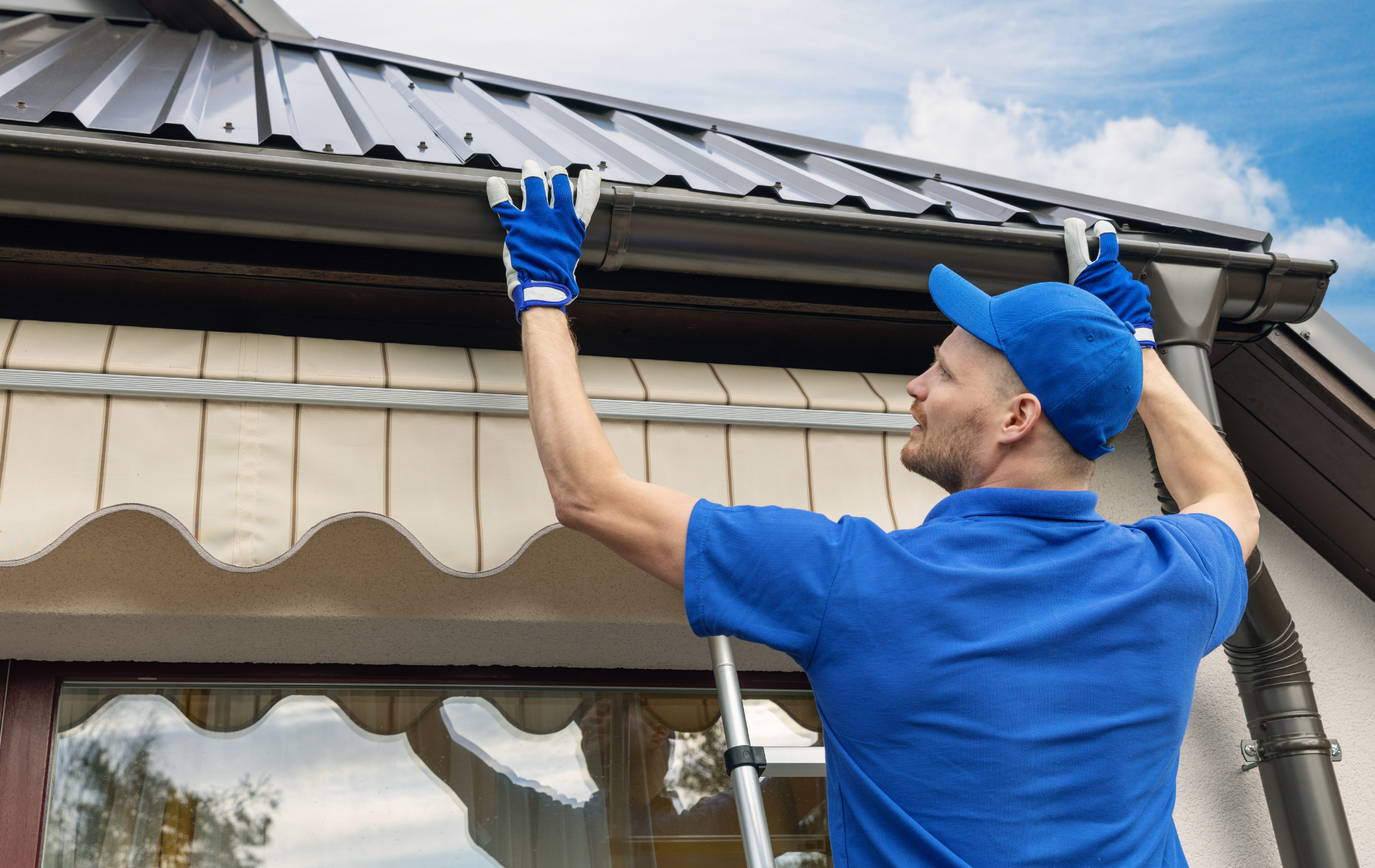 A man is standing on a ladder fixing a gutter on a roof.