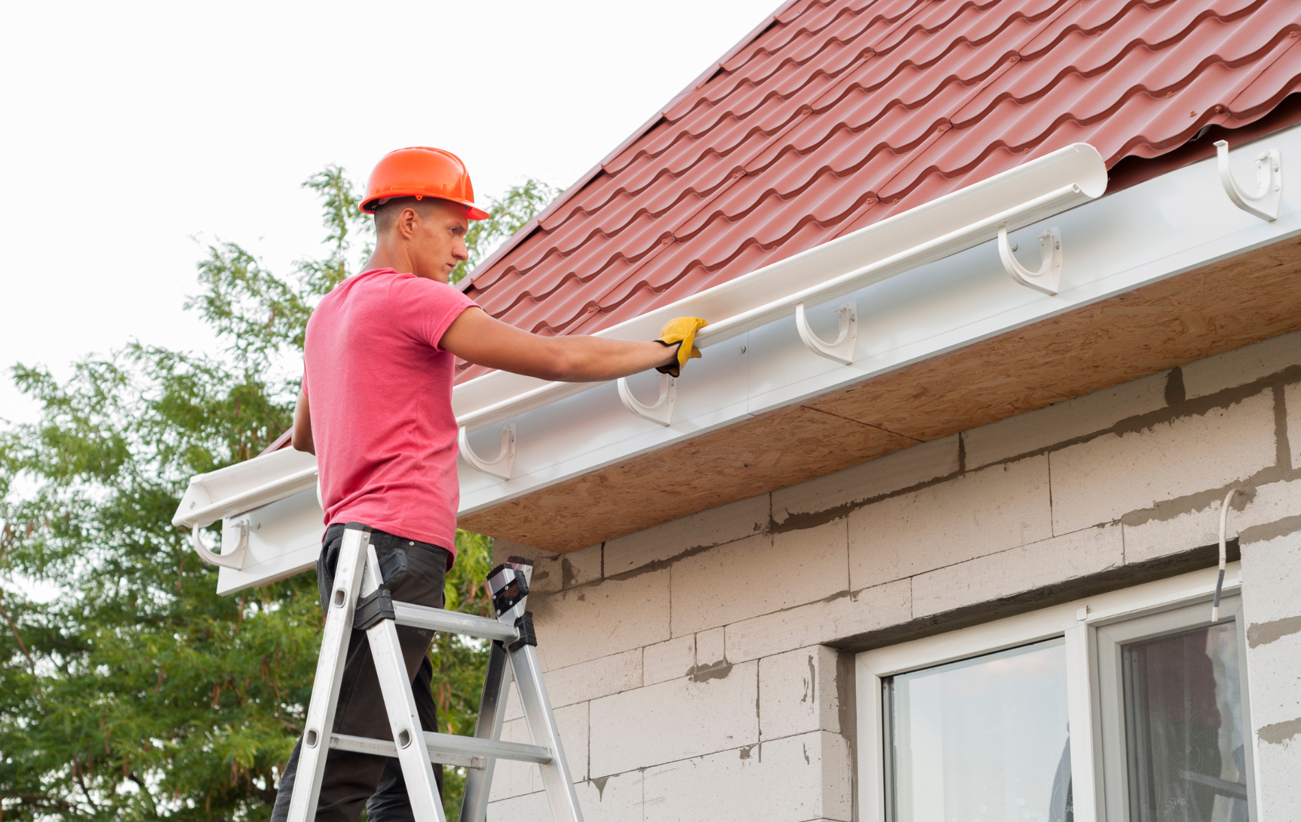 A man is standing on a ladder fixing a gutter on the side of a house.