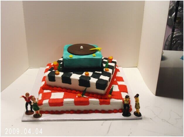 Three-tiered cake with a red and white checkered base, a black and white checkered layer, and a teal record player on top; small figurines around the base.