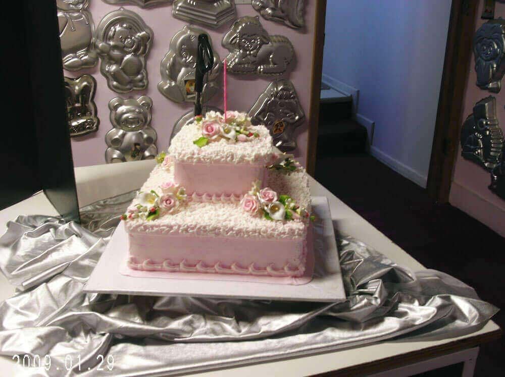 Two-tiered pink frosted cake with floral decorations on a white square serving board. Silver fabric on table.
