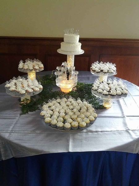 Dessert table with tiered cake, cupcakes, and cookies on glass stands, draped in white and blue.