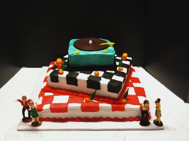 Three-tiered cake with a checkered dance floor design and record player on top; small figures dance nearby.