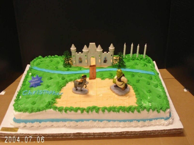 Shrek cake featuring figures of Shrek and Donkey, castle, and candles. Green frosting, blue river.