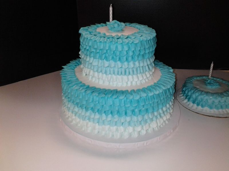 Two-tiered cake with blue and white frosting in a ruffled pattern, topped with candles. Small matching cake beside it.
