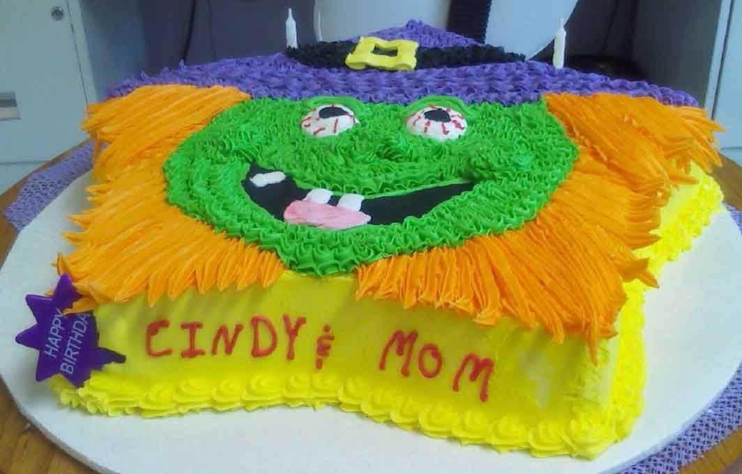 Witch-shaped birthday cake: green face, orange hair, purple hat, yellow icing with 