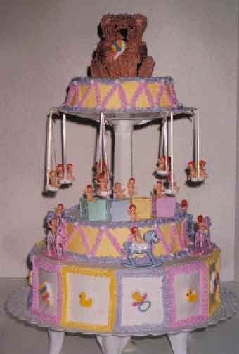 Three-tiered baby shower cake; carousel design with baby figurines and a teddy bear topper. Pastel colors.
